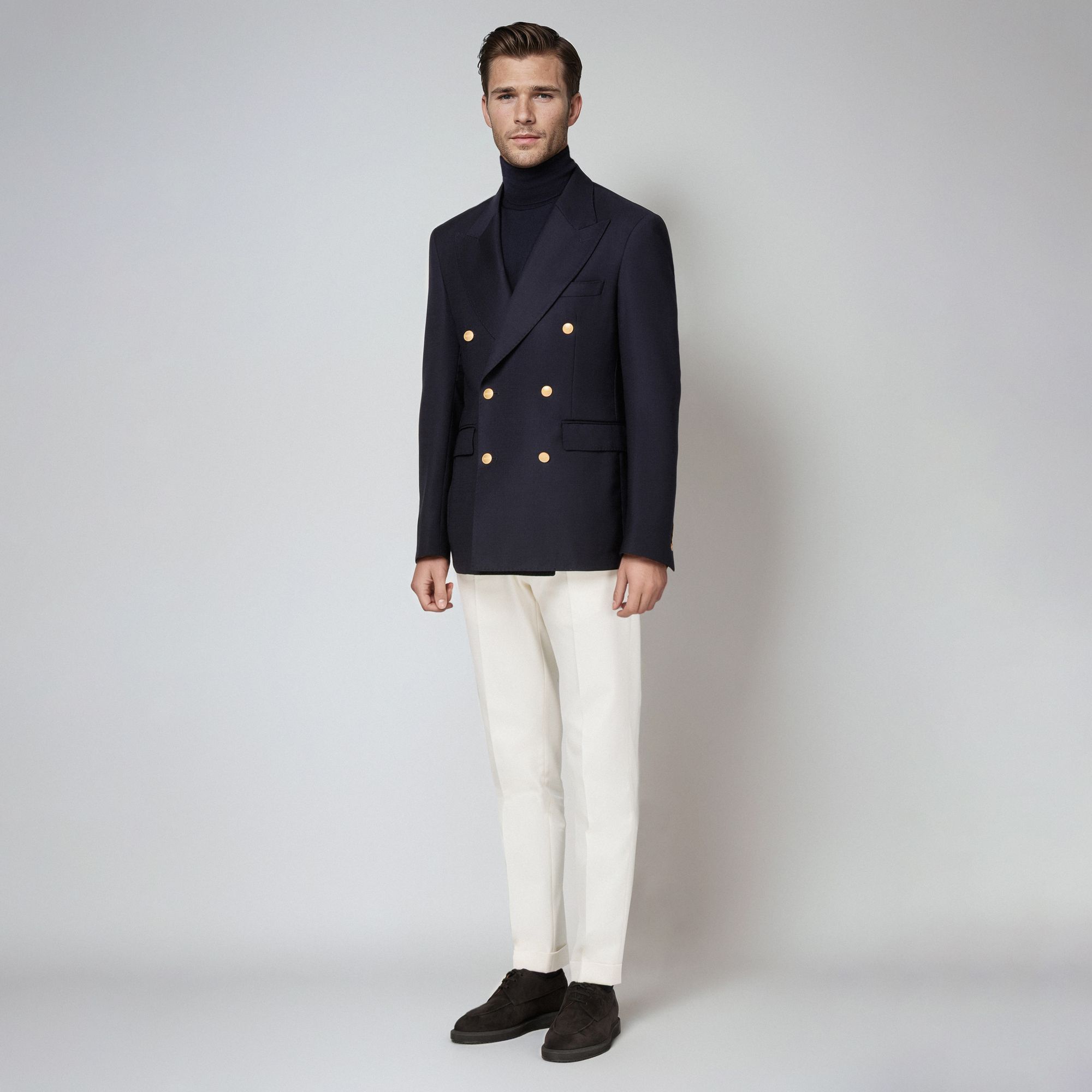 Uomo con total look Elio Evan: blazer doppiopetto blu navy con bottoni dorati, dolcevita blu e pantaloni bianchi.
